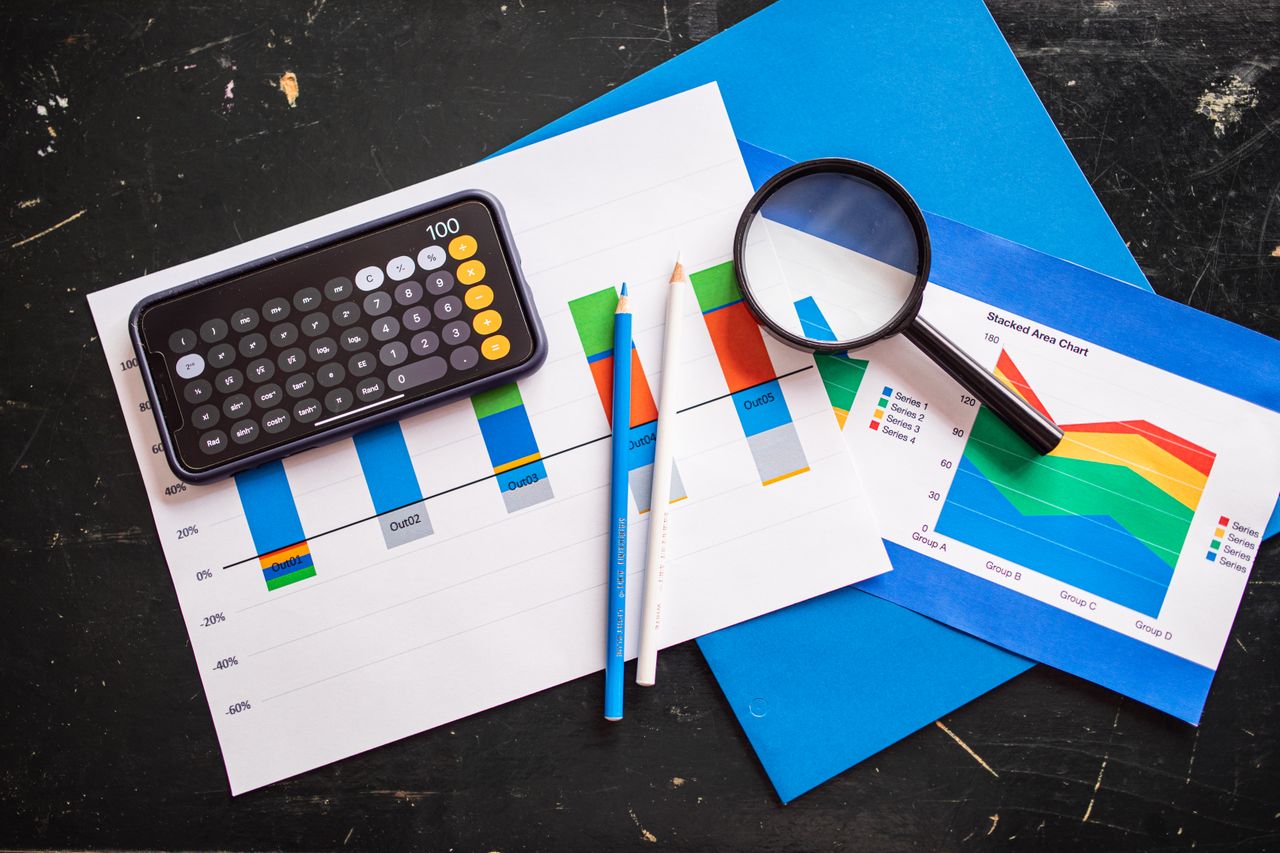 services-02 Top view of financial charts with a smartphone calculator, magnifying glass, and pencils on a desk.